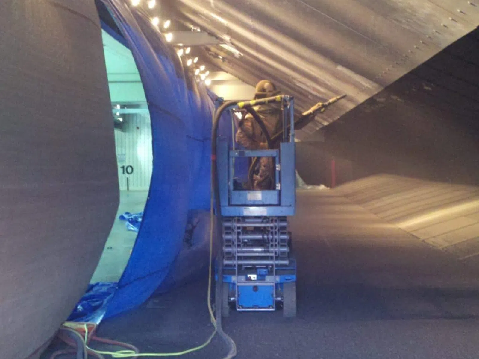 Worker on lift machine spray painting large industrial metal surface inside facility with bright overhead lights.