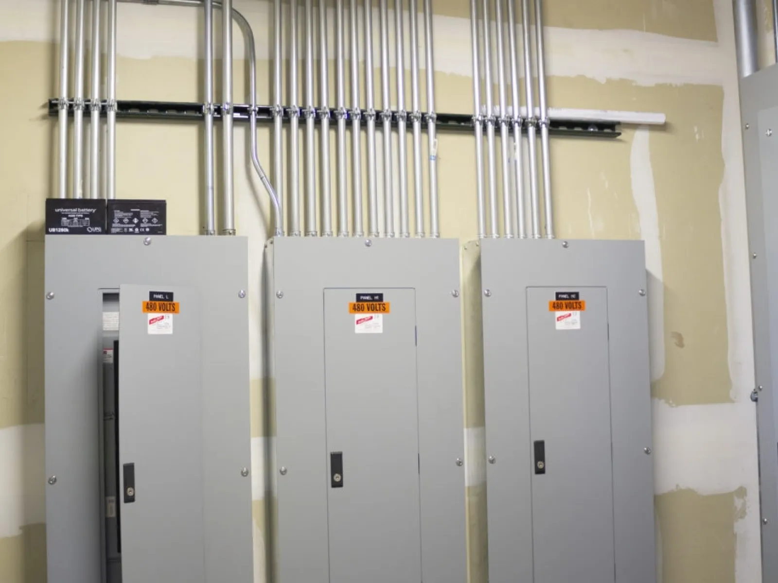 Three gray electrical panels with 480 volts caution labels and metal conduit piping above on beige wall.