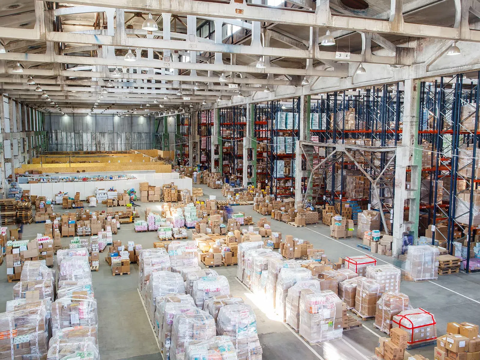 Large warehouse interior with stacked pallets, boxes, and tall metal shelving under bright lighting