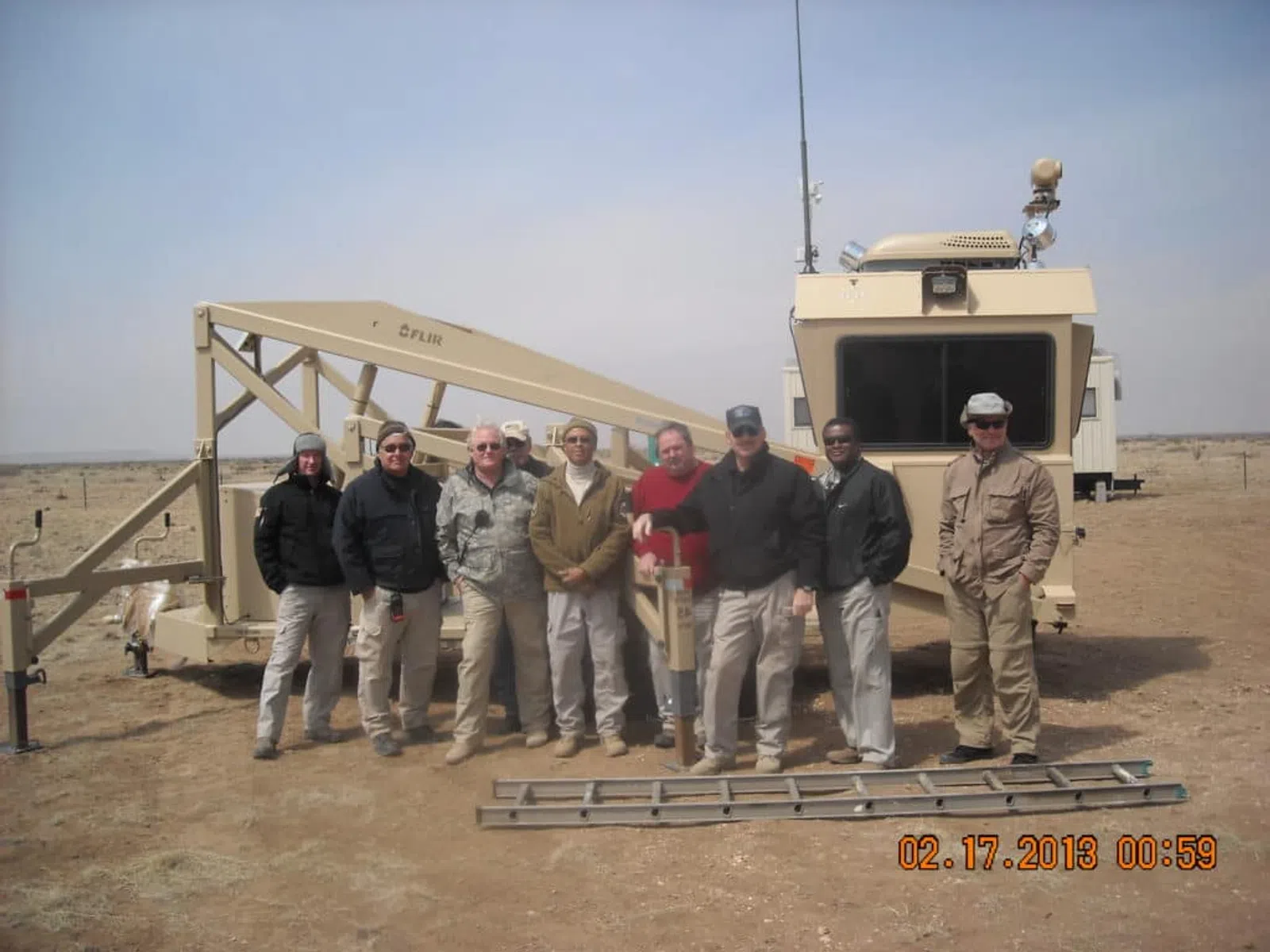 Group of eight men standing in front of a large beige military or construction vehicle in a desert landscape.