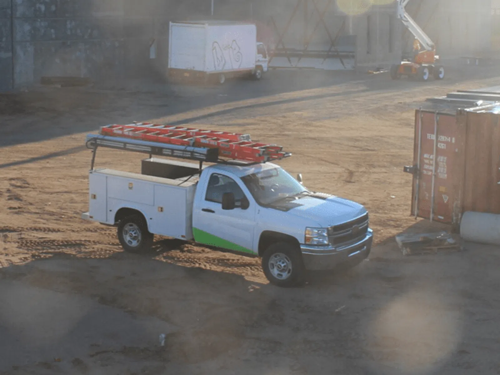 White utility truck with ladders parked on a dusty construction site near concrete buildings and a lift.