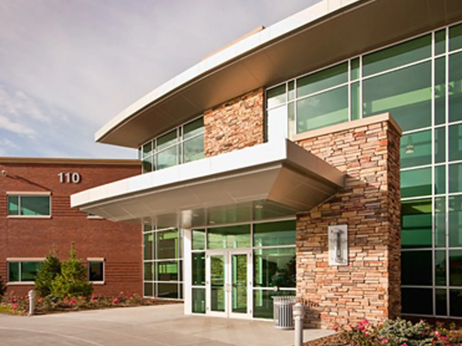 Modern commercial building entrance with glass windows, stone facade, and landscaped surroundings under a clear sky.