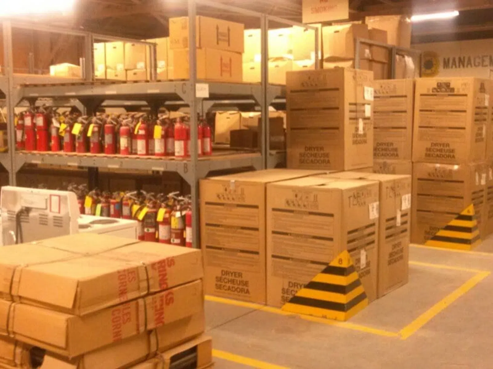 Warehouse storage with stacked cardboard boxes labeled dryer and shelves filled with multiple fire extinguishers.