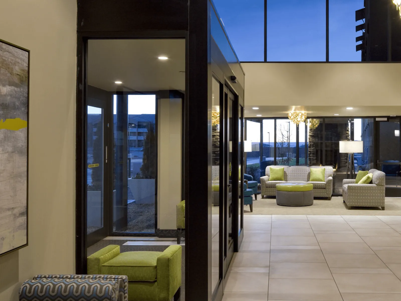 Modern hotel lobby with green and gray armchairs, large windows, and contemporary lighting at dusk.