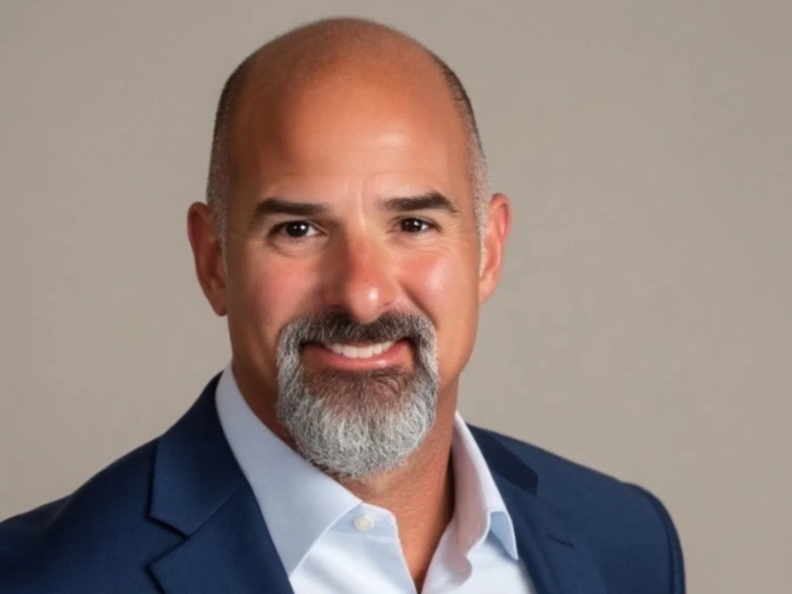 Smiling bald man with a gray beard wearing a blue suit jacket and white shirt against a plain background