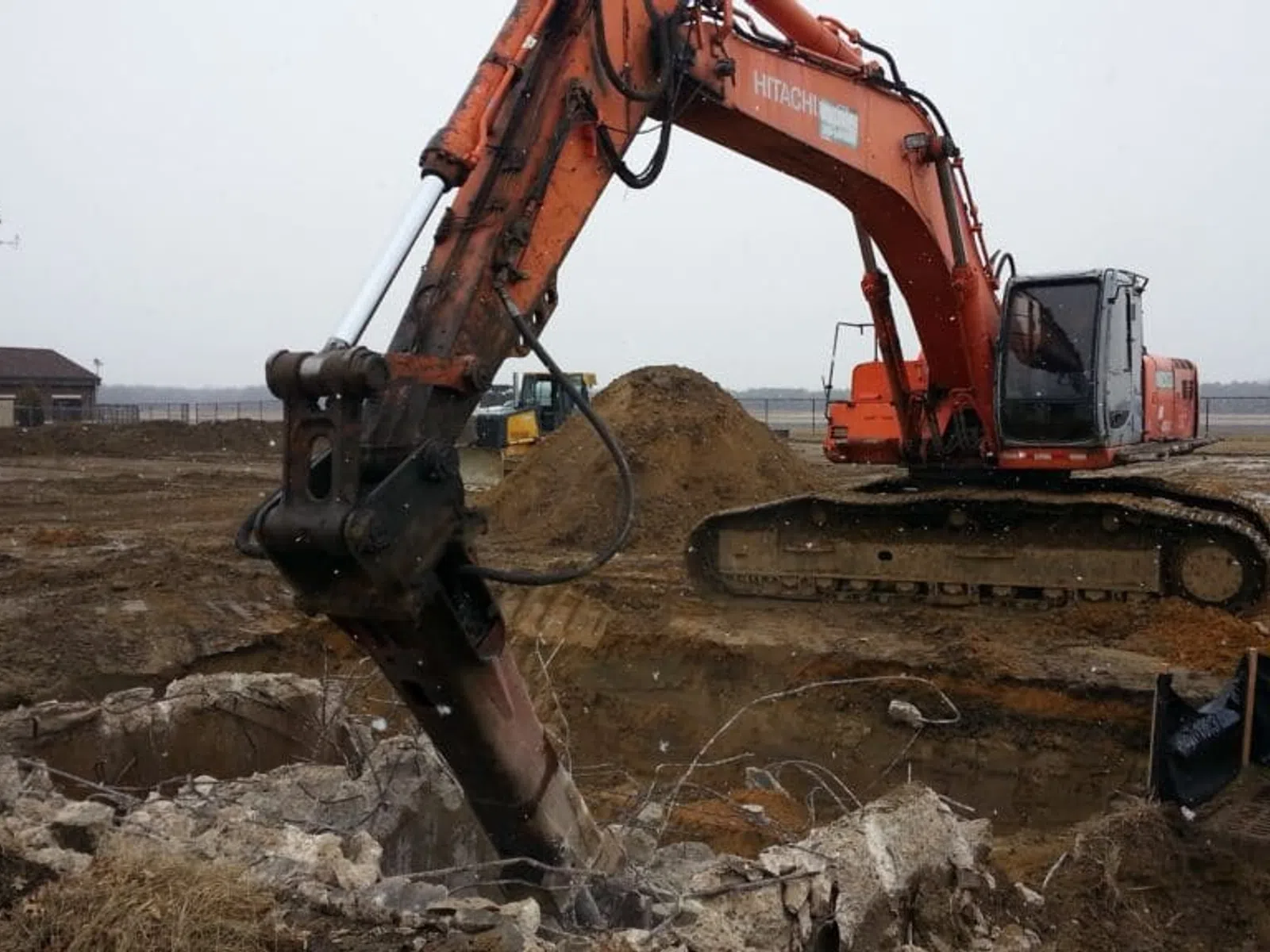Orange excavator with hydraulic hammer breaking concrete at a construction site with dirt and debris around.