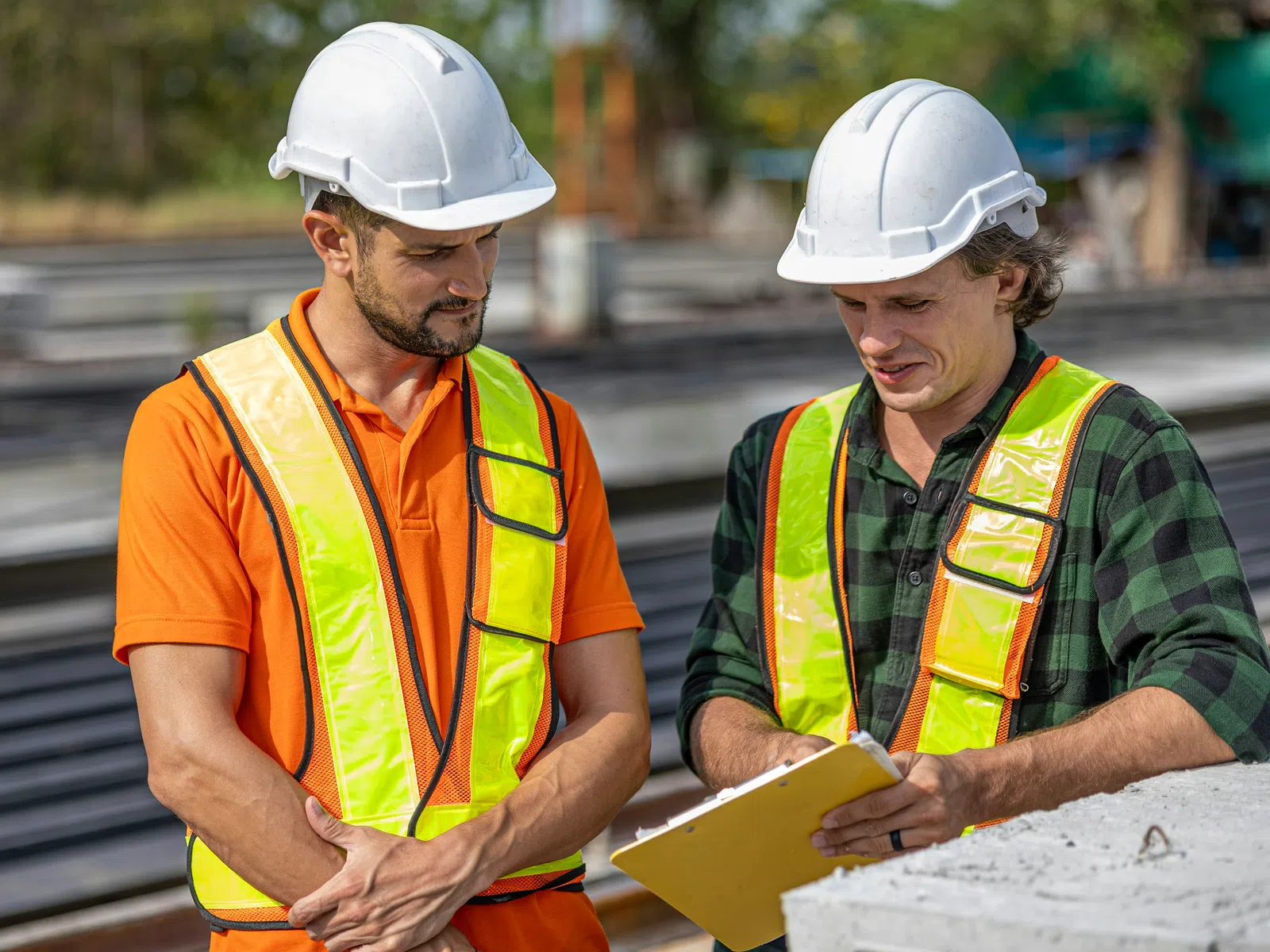 Two construction workers wearing white helmets and reflective vests reviewing plans on a clipboard outdoors.