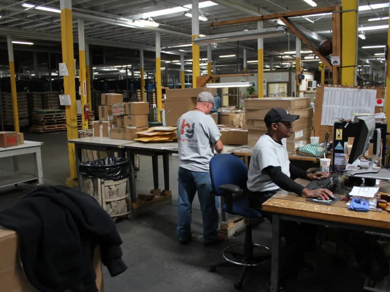 Two workers managing packages and using a computer in a large warehouse with stacked boxes and industrial lighting.