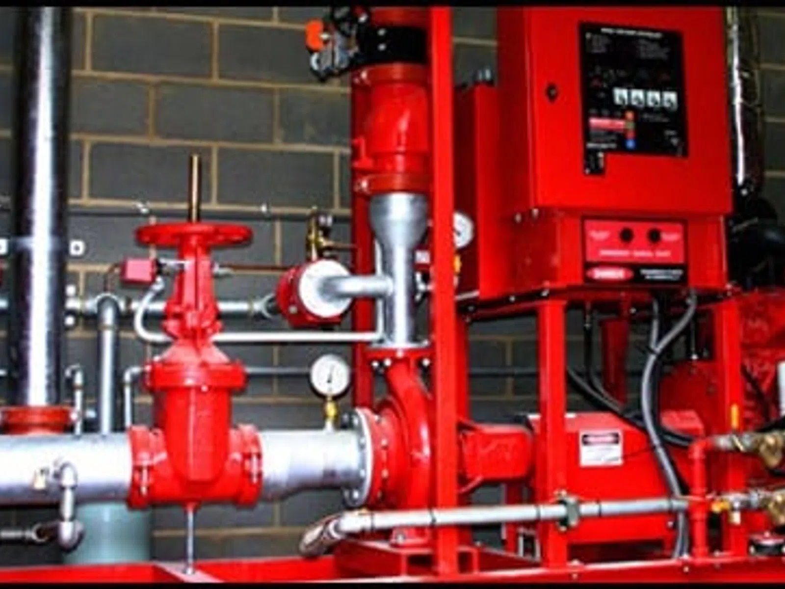 Industrial red fire sprinkler system with valves, gauges, and control panel mounted on gray brick wall