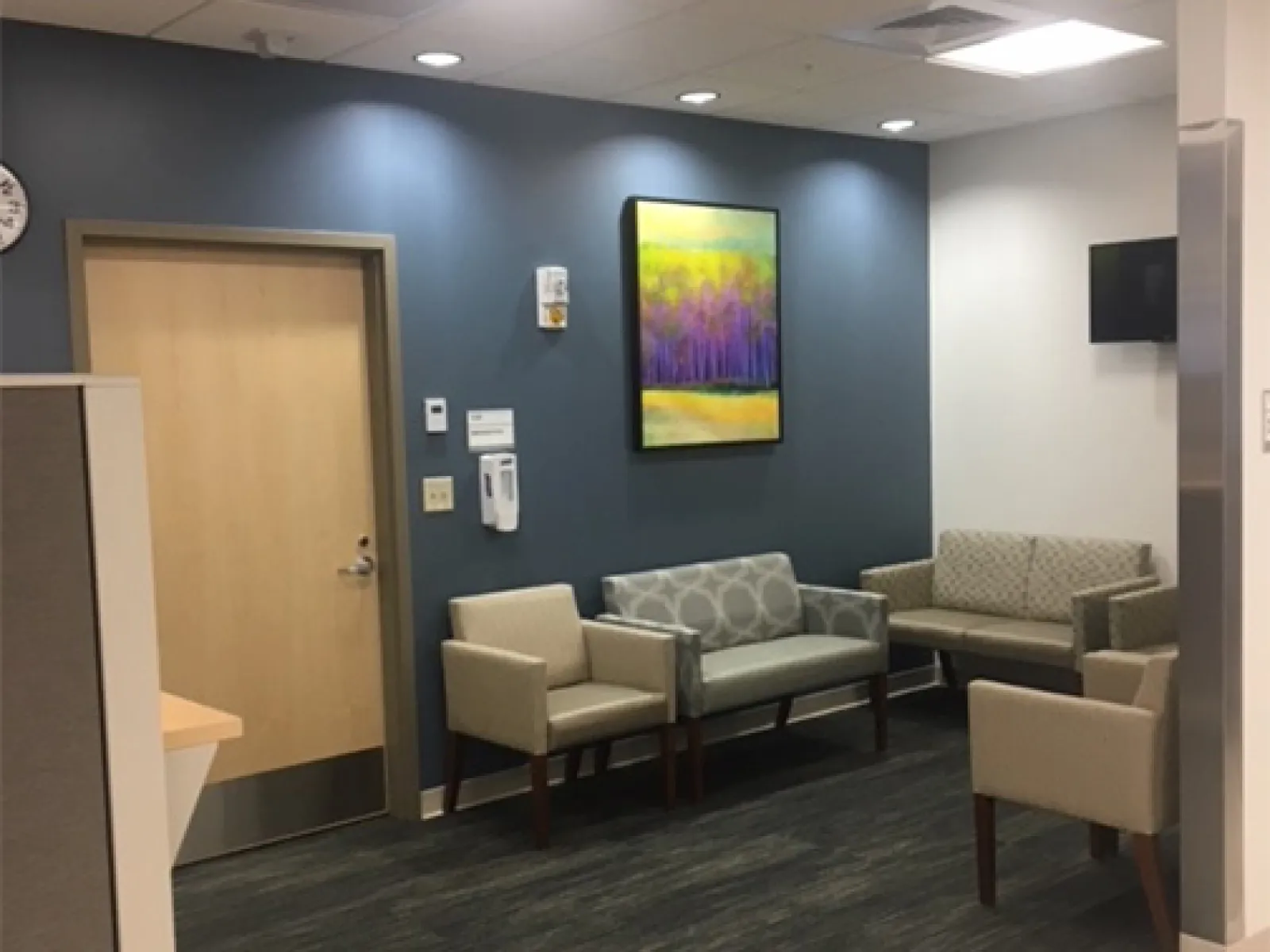 Modern waiting area with beige chairs, gray-patterned loveseat, dark wood floor, blue accent wall, and colorful painting.