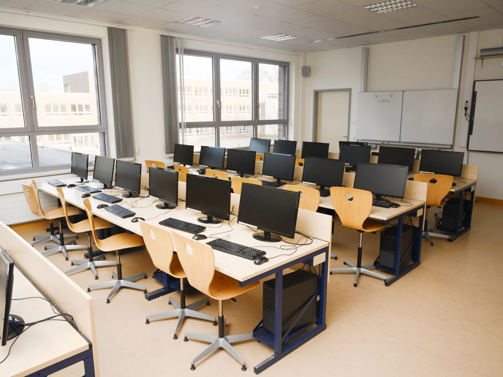 Empty computer lab with multiple desktop computers, wooden chairs, large windows, and whiteboards in a classroom.