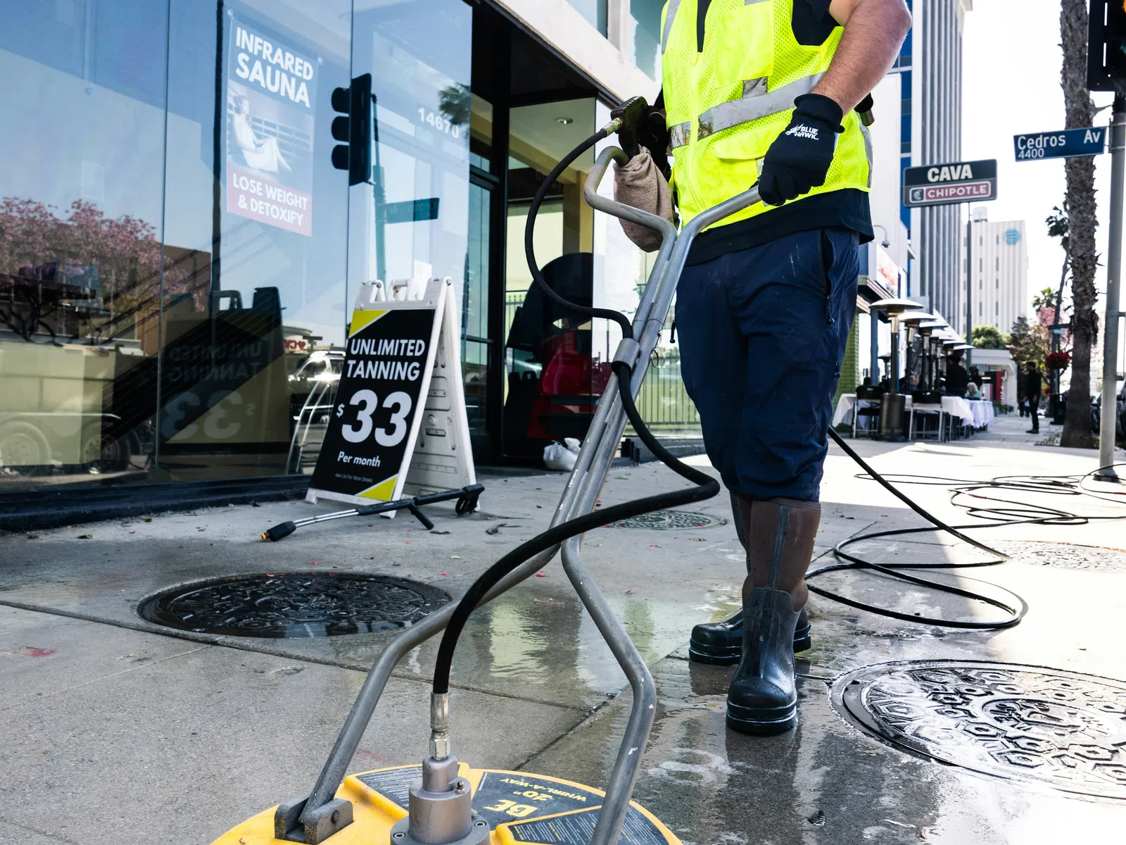 Worker in reflective vest power washing sidewalk with yellow surface cleaner machine in urban area
