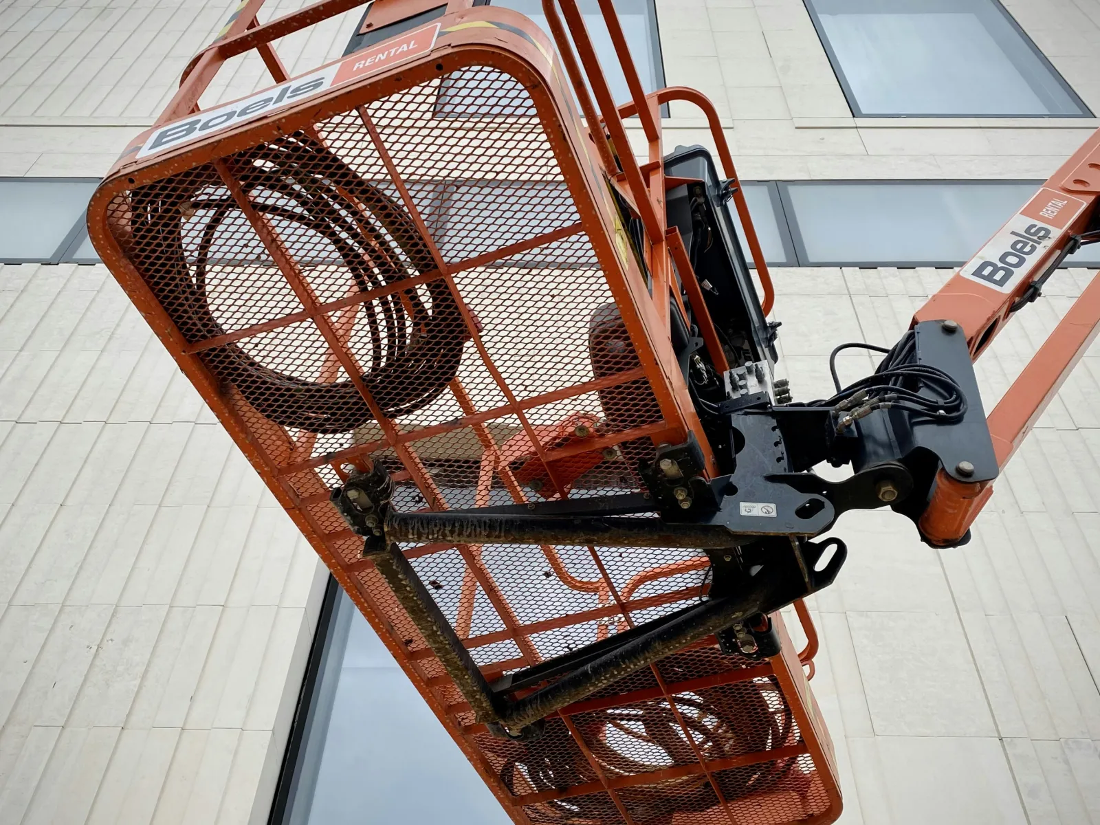 Orange Boels rented JLG lift platform extended upward in front of a modern building facade.