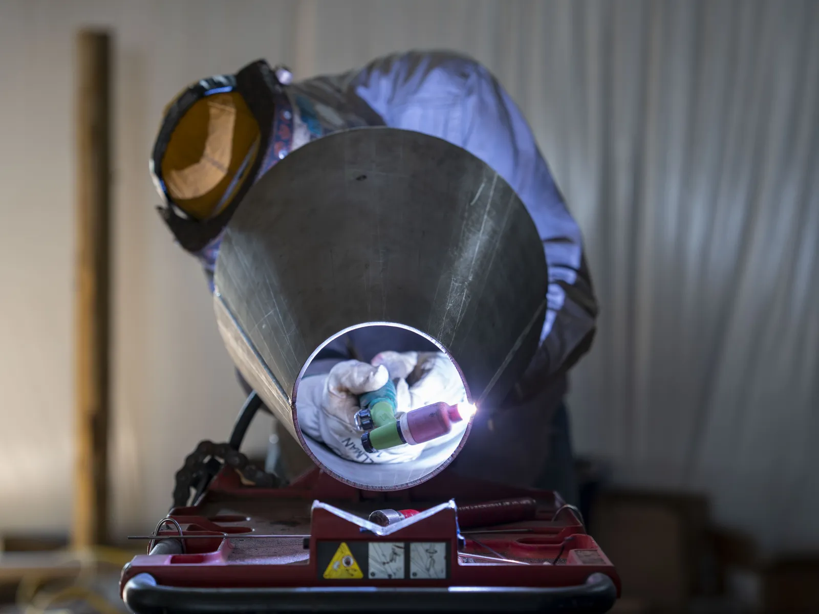 Welder in protective gear welding inside a large metal pipe with bright sparks visible through the pipe opening