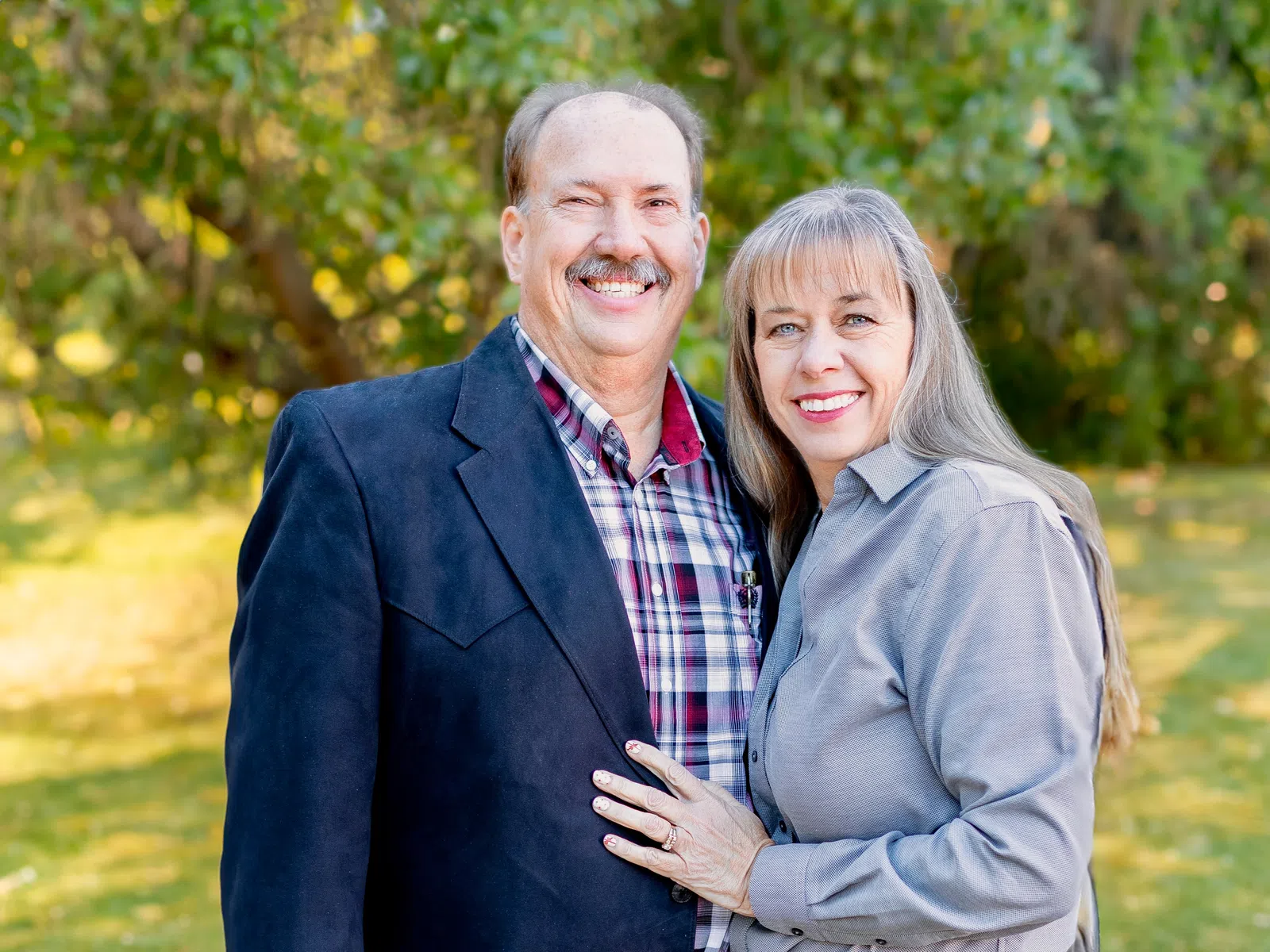 Smiling middle-aged couple outdoors in casual and formal attire with green trees and grass background.