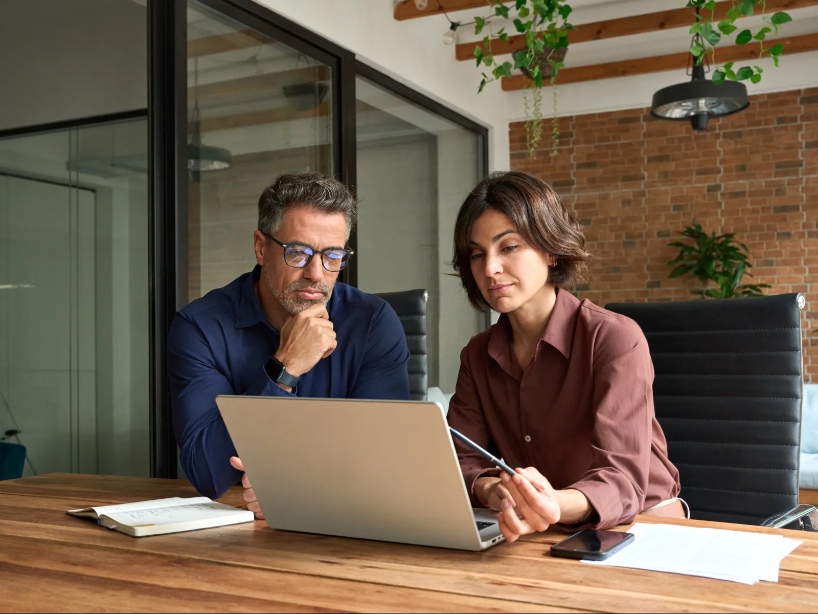 Two professionals collaborating and reviewing content on a laptop in a modern office space with wooden beams and plants.