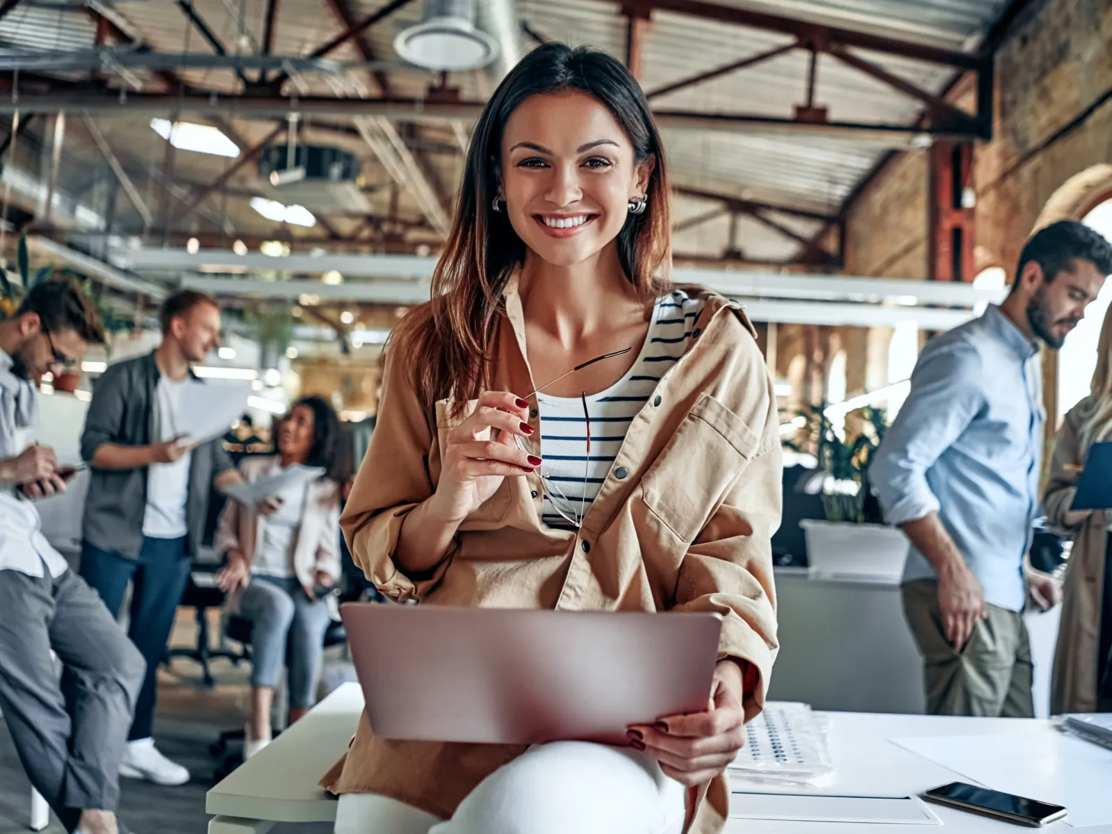 Smiling woman with laptop and glasses in modern office with colleagues collaborating in background