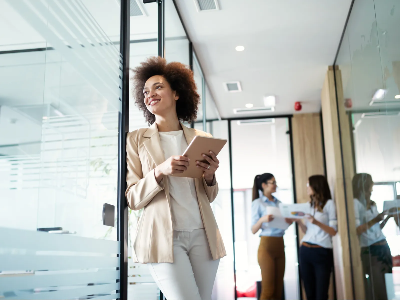 Confident businesswoman holding a tablet while colleagues discuss documents in a modern office hallway