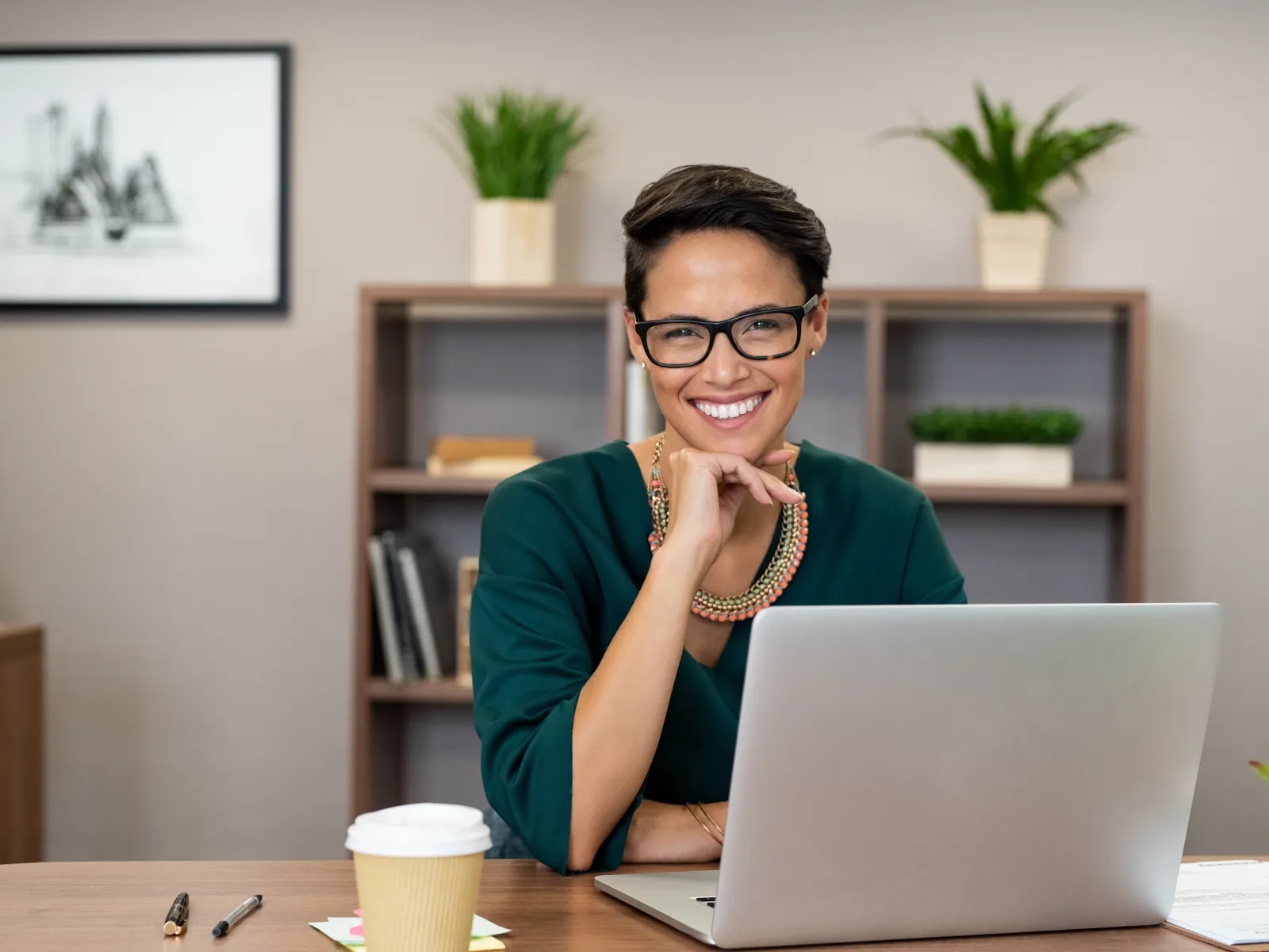 Smiling businesswoman with glasses sitting at desk with laptop in modern office space.