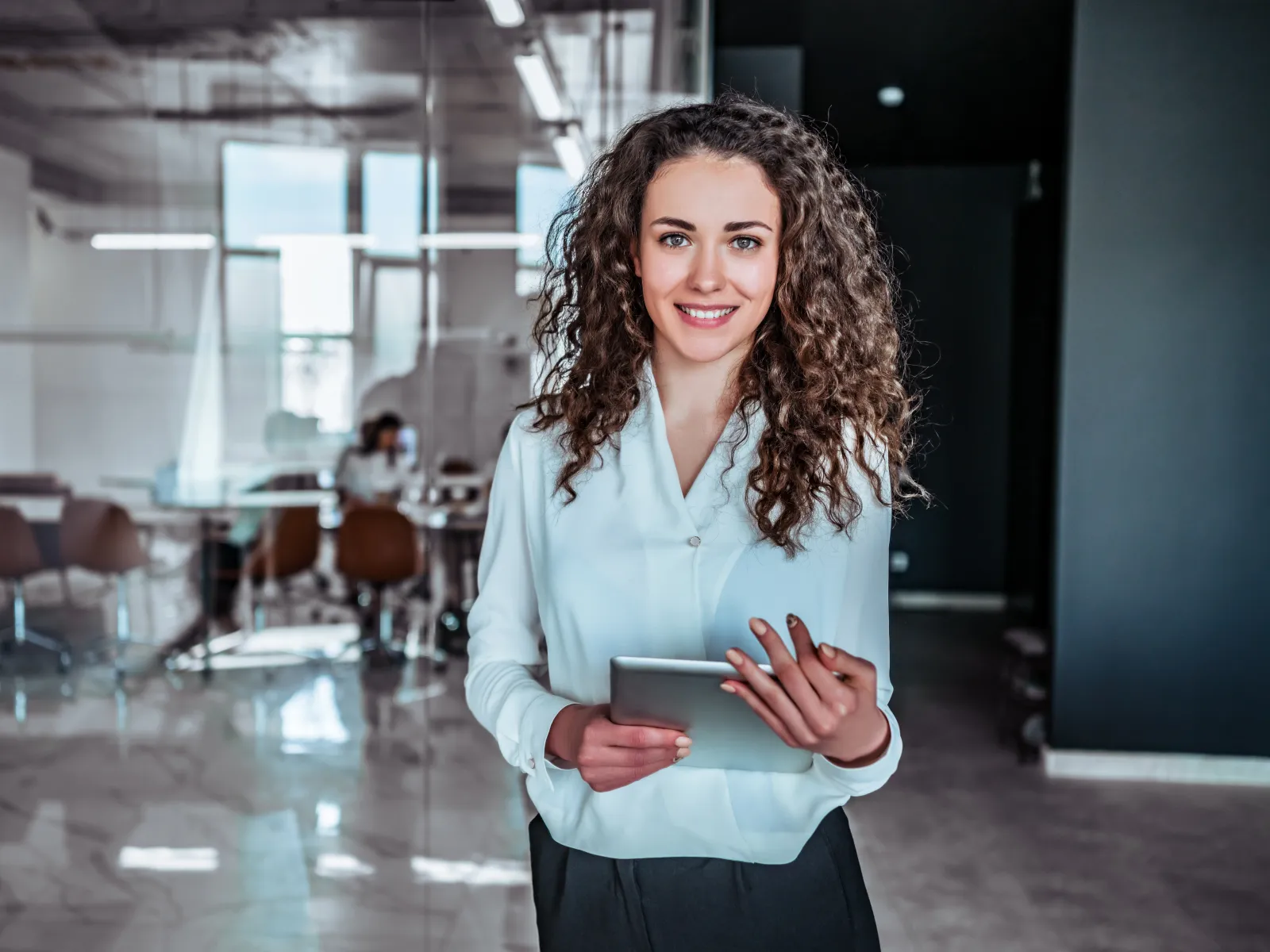 Smiling businesswoman holding a tablet in a modern office with glass walls and coworking space in background