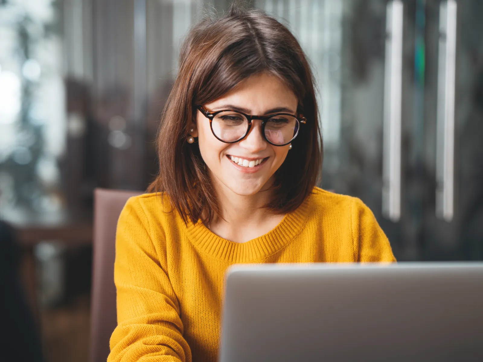 Smiling young woman in glasses and yellow sweater working on a laptop in a modern office setting