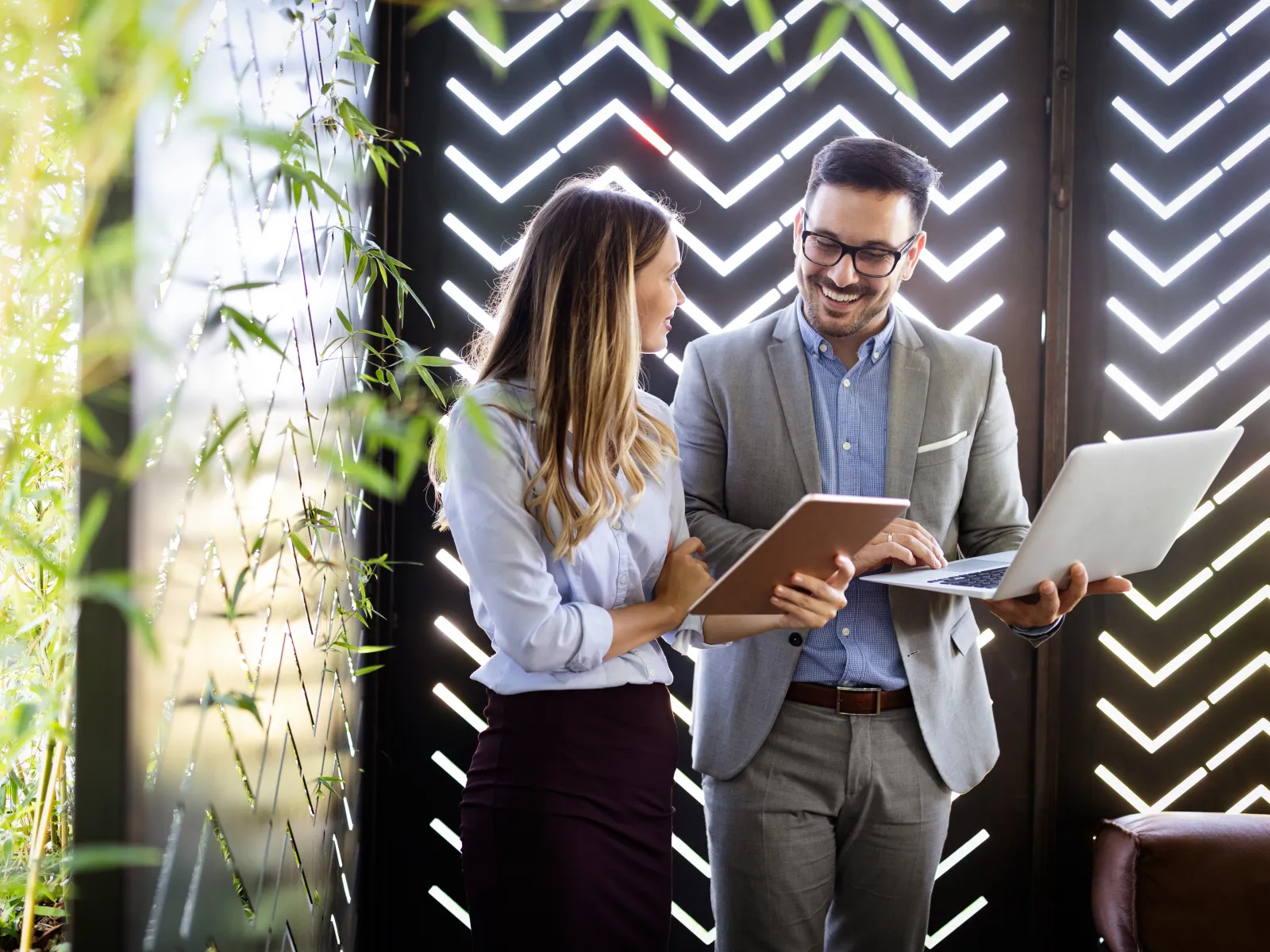 Two professionals discuss work using tablet and laptop in modern office with geometric light wall and greenery.