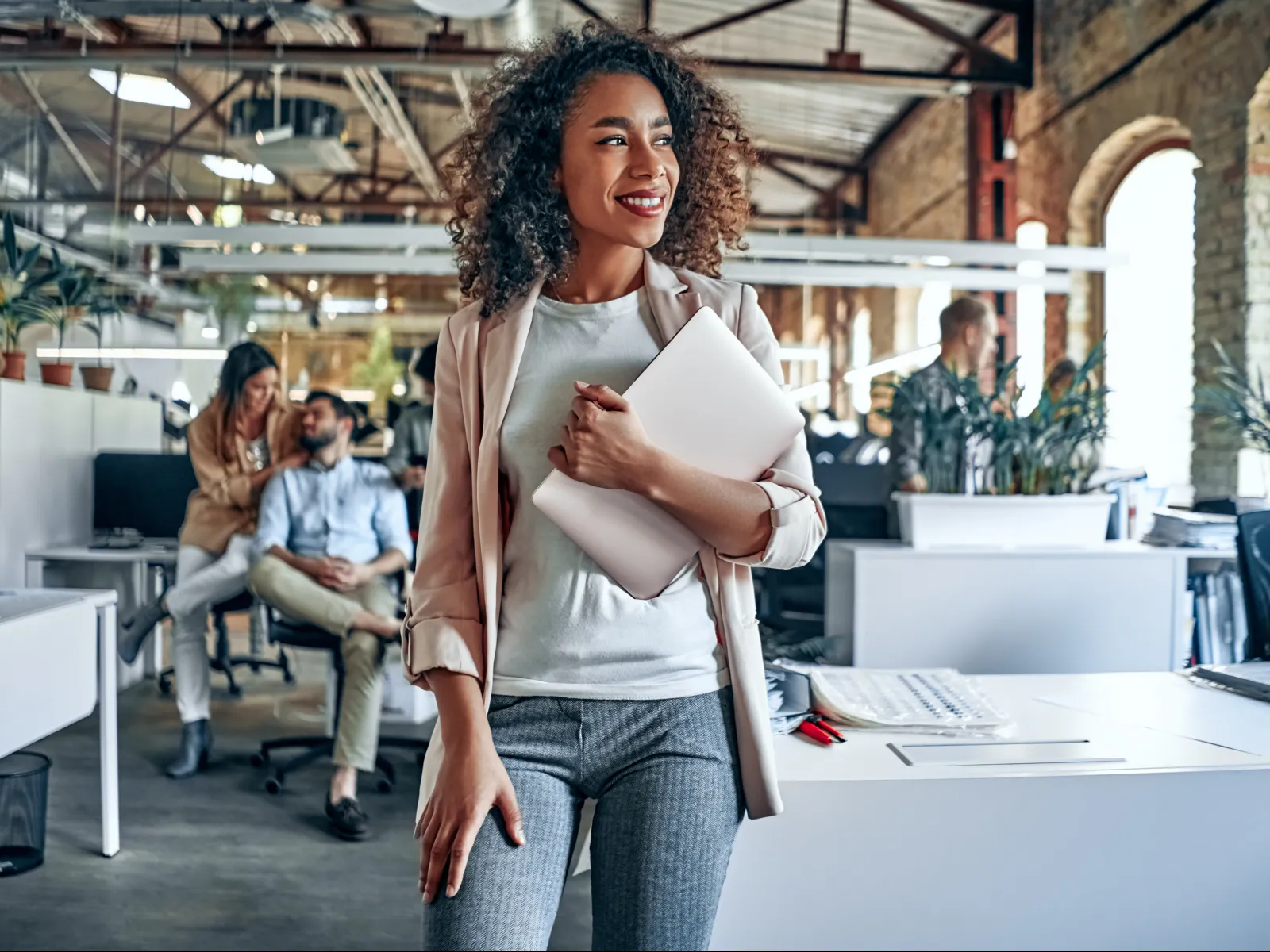 Smiling woman holding laptop in modern office with colleagues and large windows in background