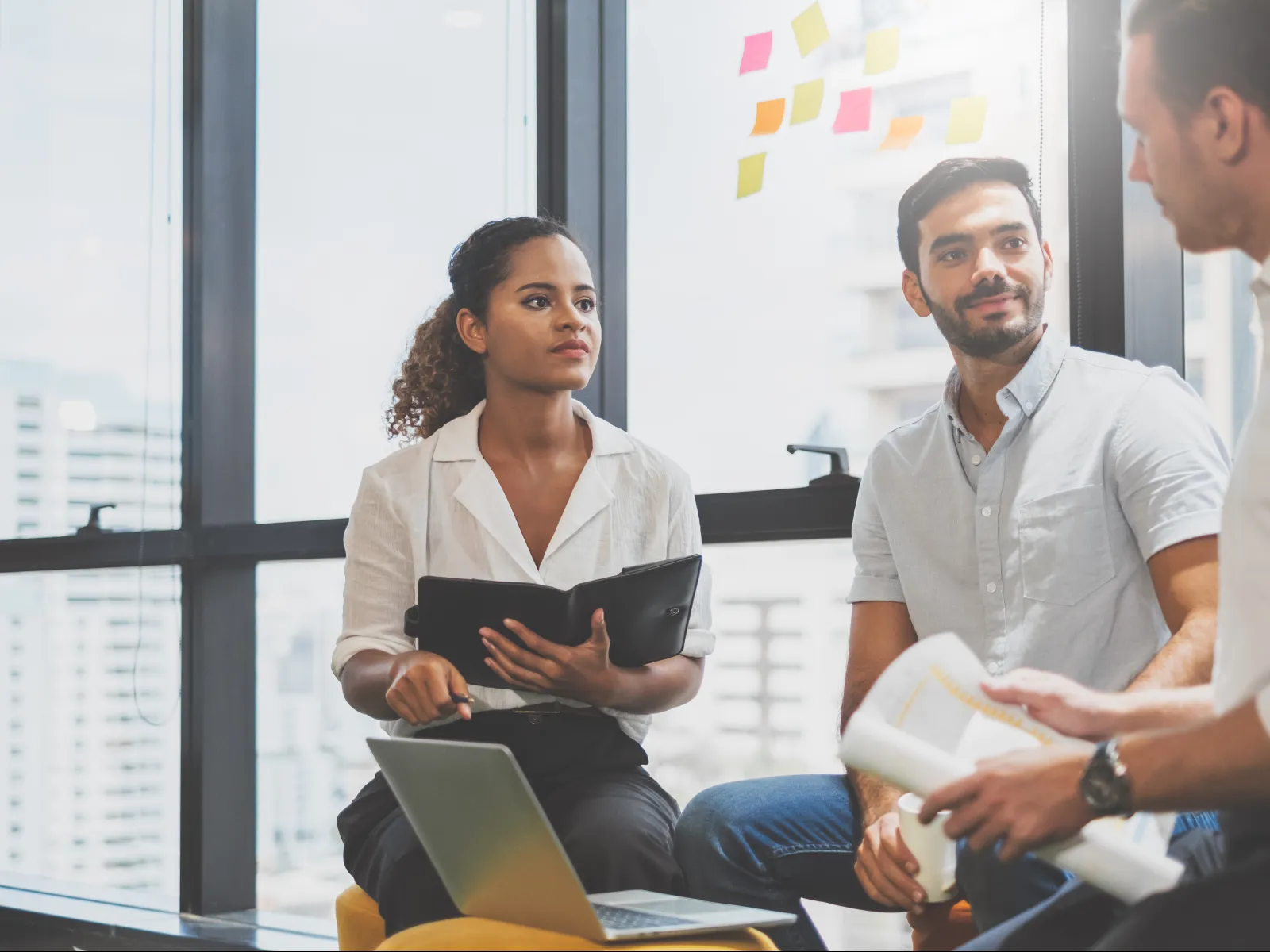 Three coworkers engaged in a meeting with documents, laptop, and sticky notes in a bright office space.