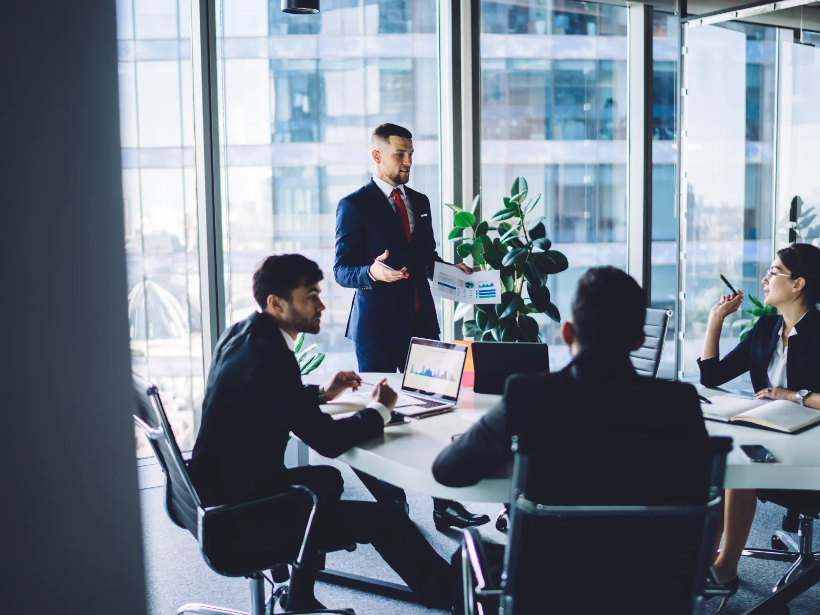 Business team in formal attire discussing charts and data during a meeting in a modern glass office.