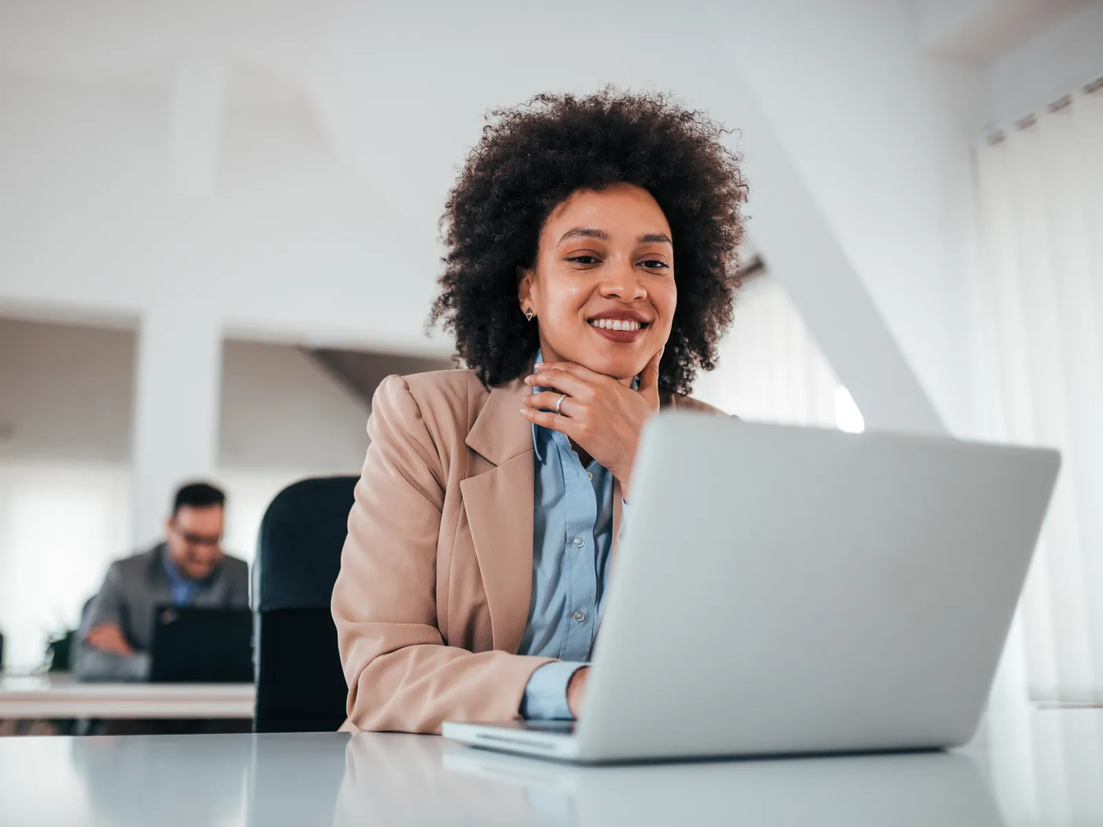 Professional woman with curly hair working on laptop in bright modern office setting with colleague in background