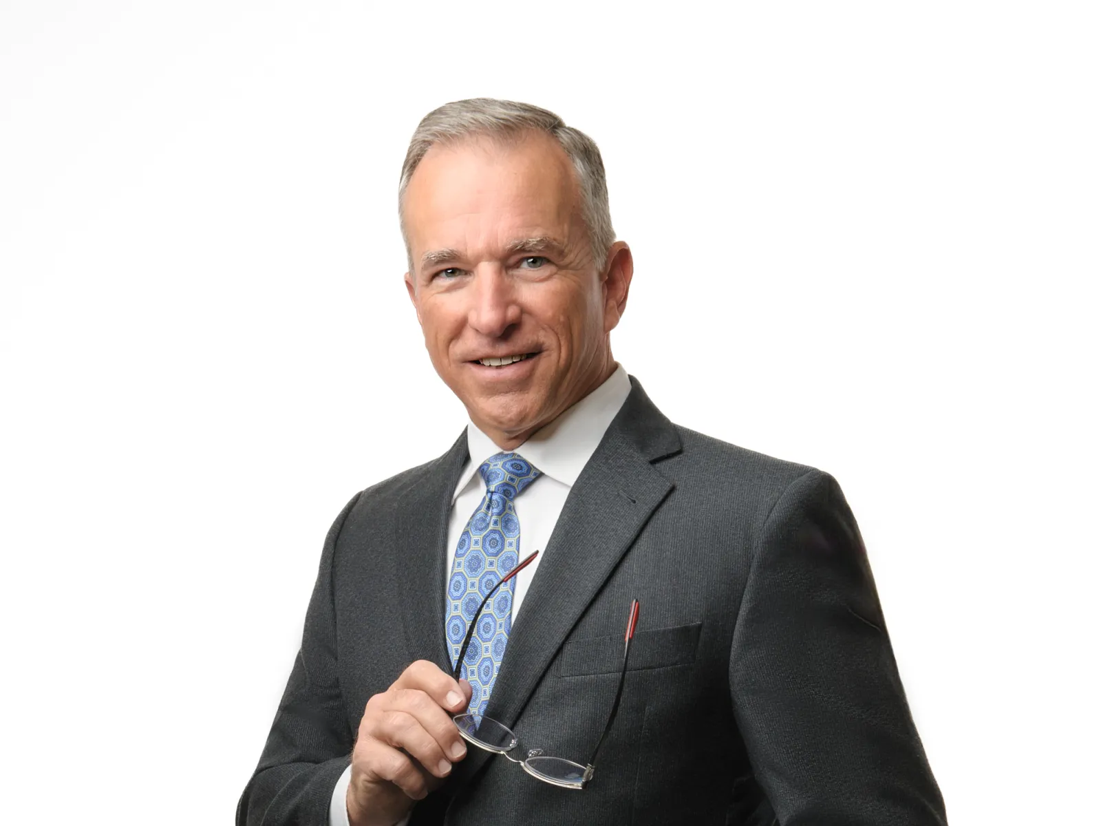 Mature businessman in dark suit and patterned blue tie holding glasses, smiling confidently against white background