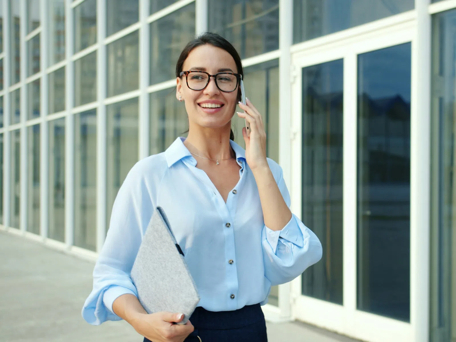 Smiling businesswoman in glasses holding folder and talking on phone outside modern office building.