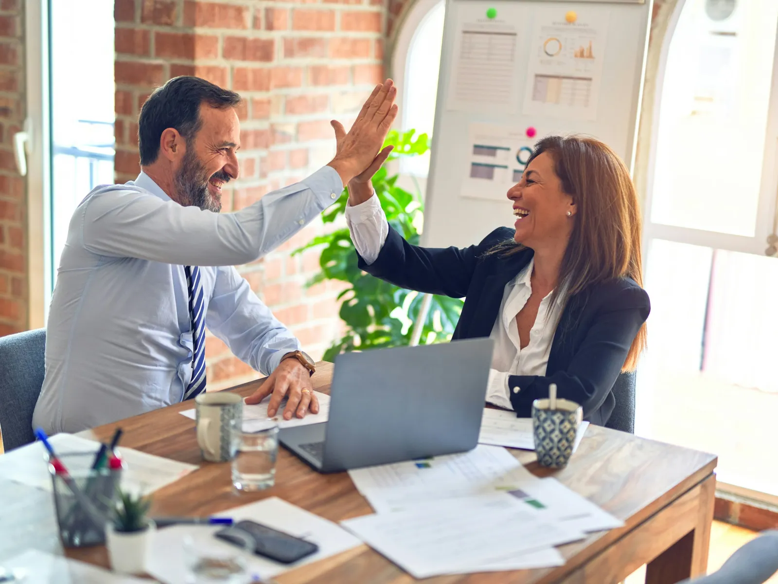 Two business colleagues in office celebrating success with a high five at a desk with laptop and documents