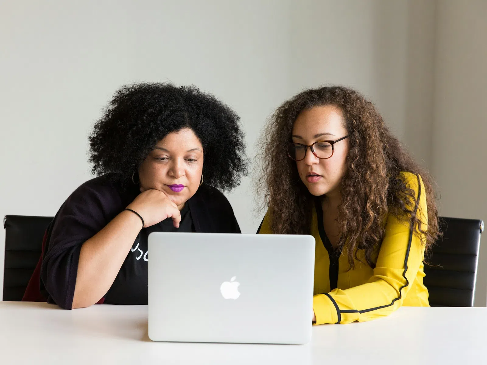 Two women focused on a MacBook laptop screen during a work meeting at a white table.