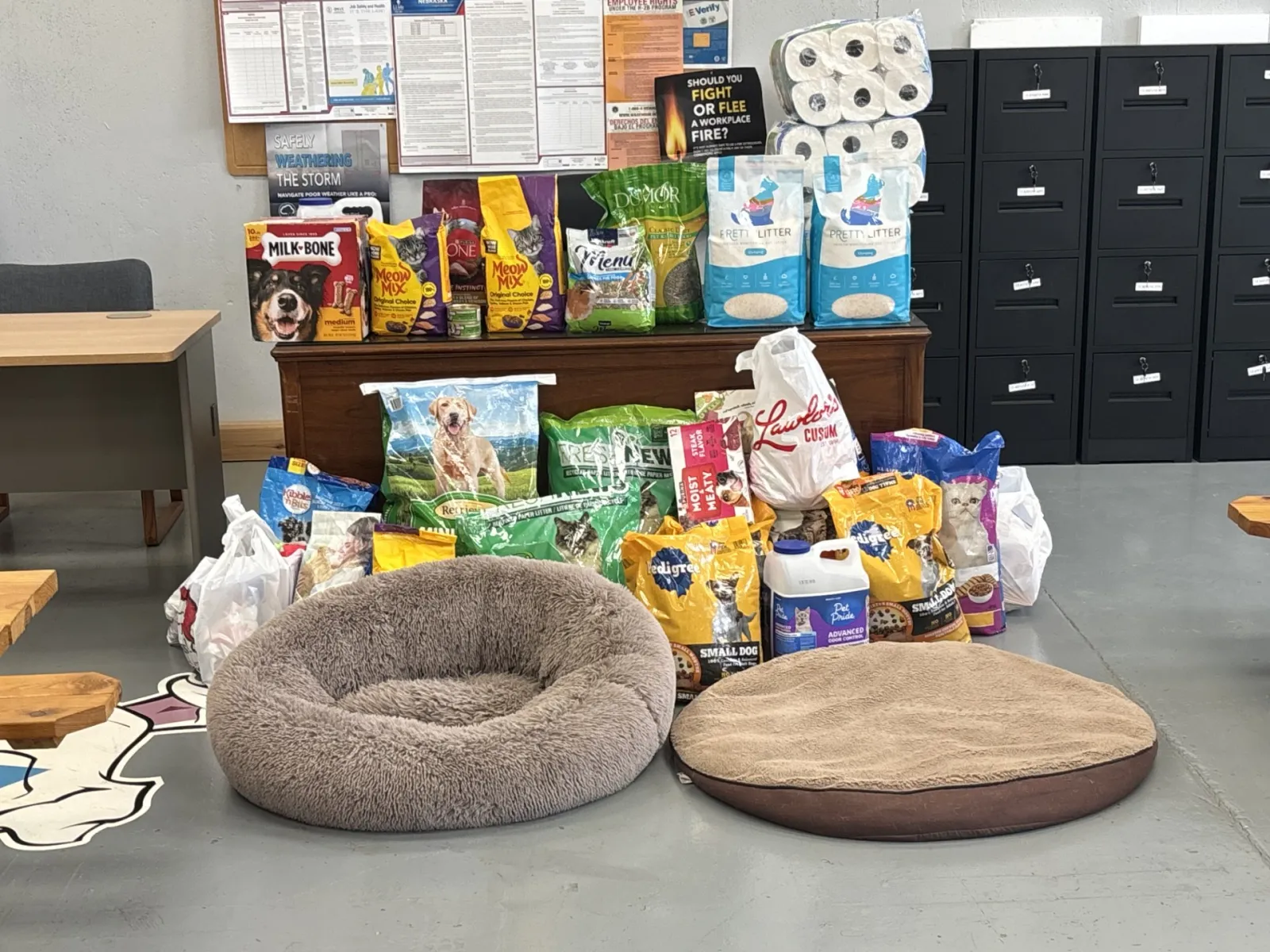 Assortment of pet supplies including dog food, cat food, litter, treats, and two pet beds arranged on a floor