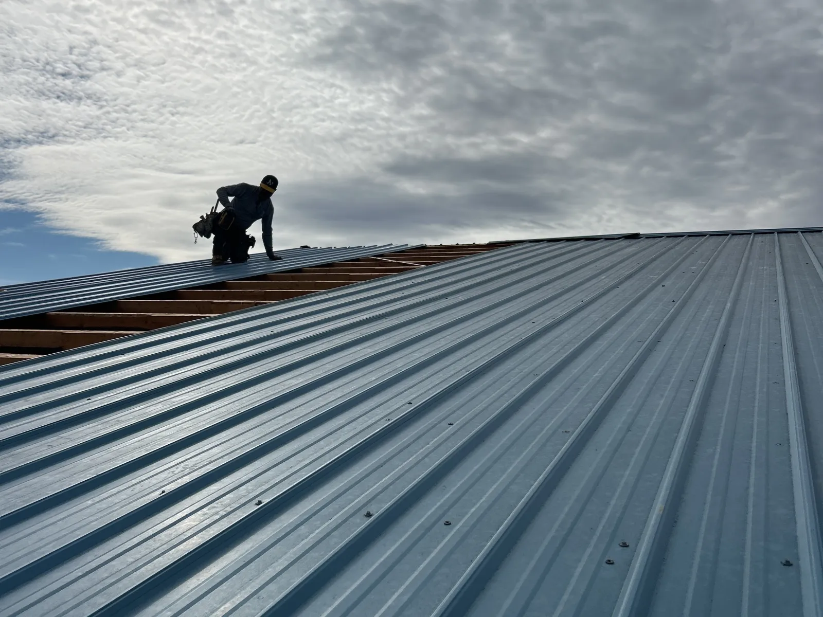 Worker installing metal roofing panels on a building under cloudy sky with sun behind clouds