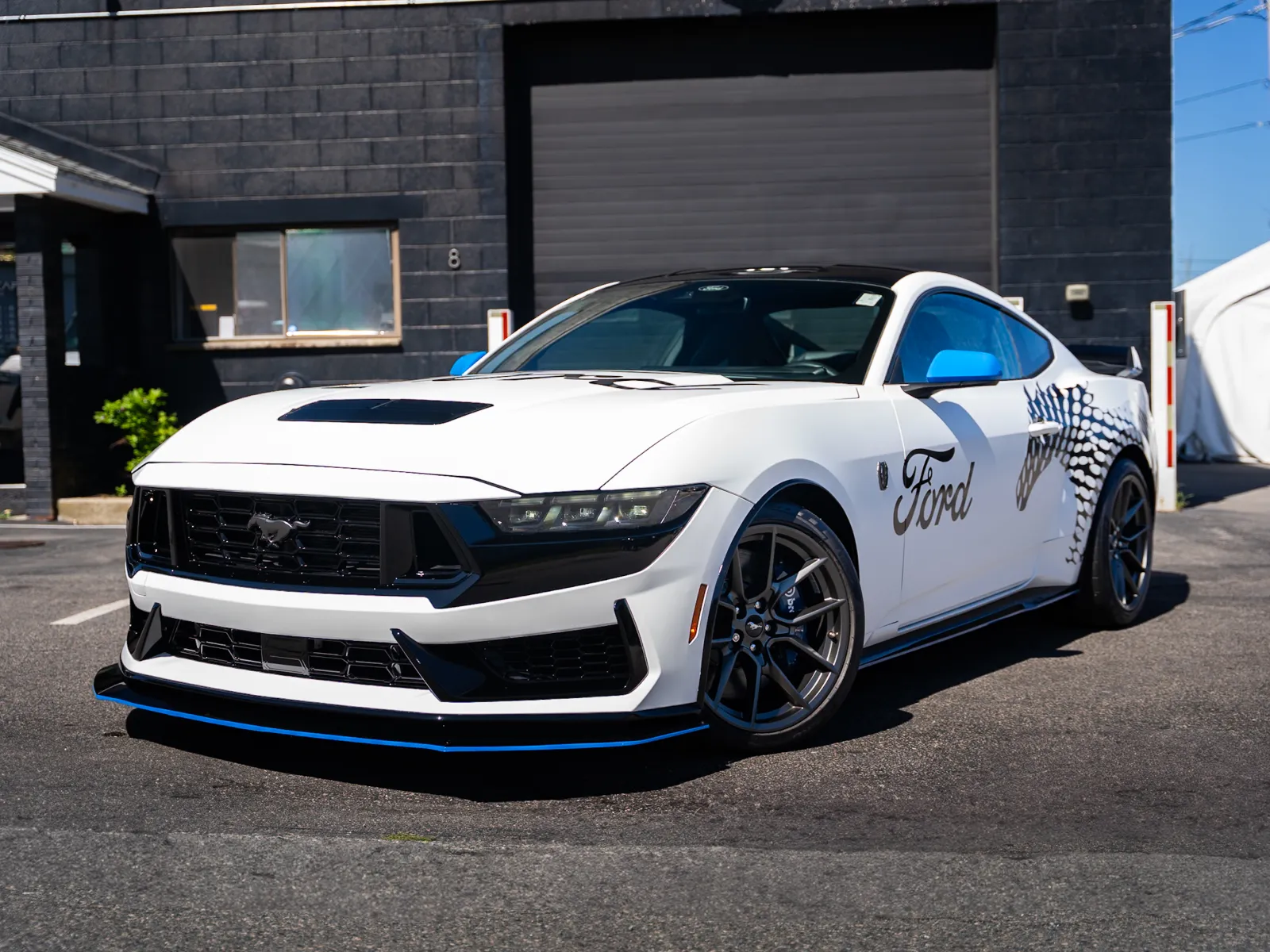 White Ford Mustang sports car with black and blue accents parked in front of a black garage door on sunny day