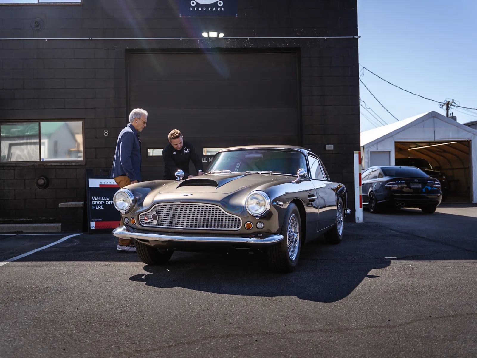 Classic vintage car parked outside a garage with two men inspecting the vehicle under sunlight.