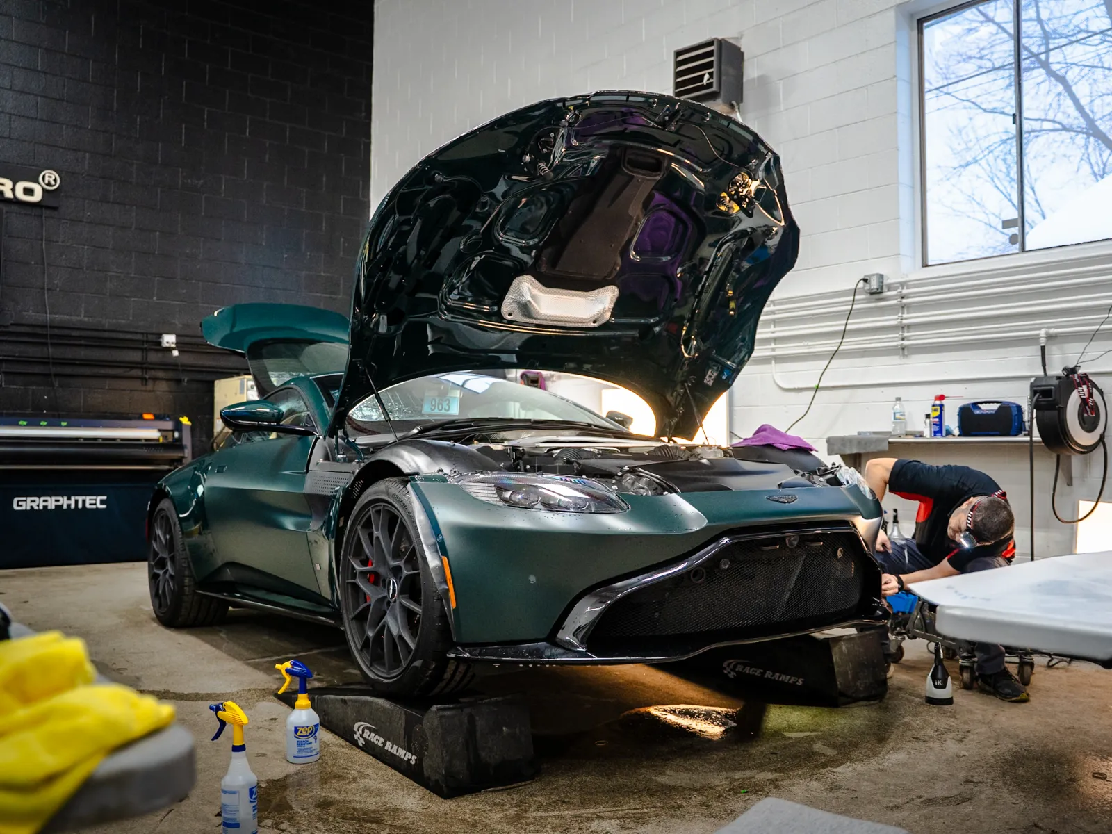 Green sports car with open hood in garage being serviced by mechanic with tools under bright window light.