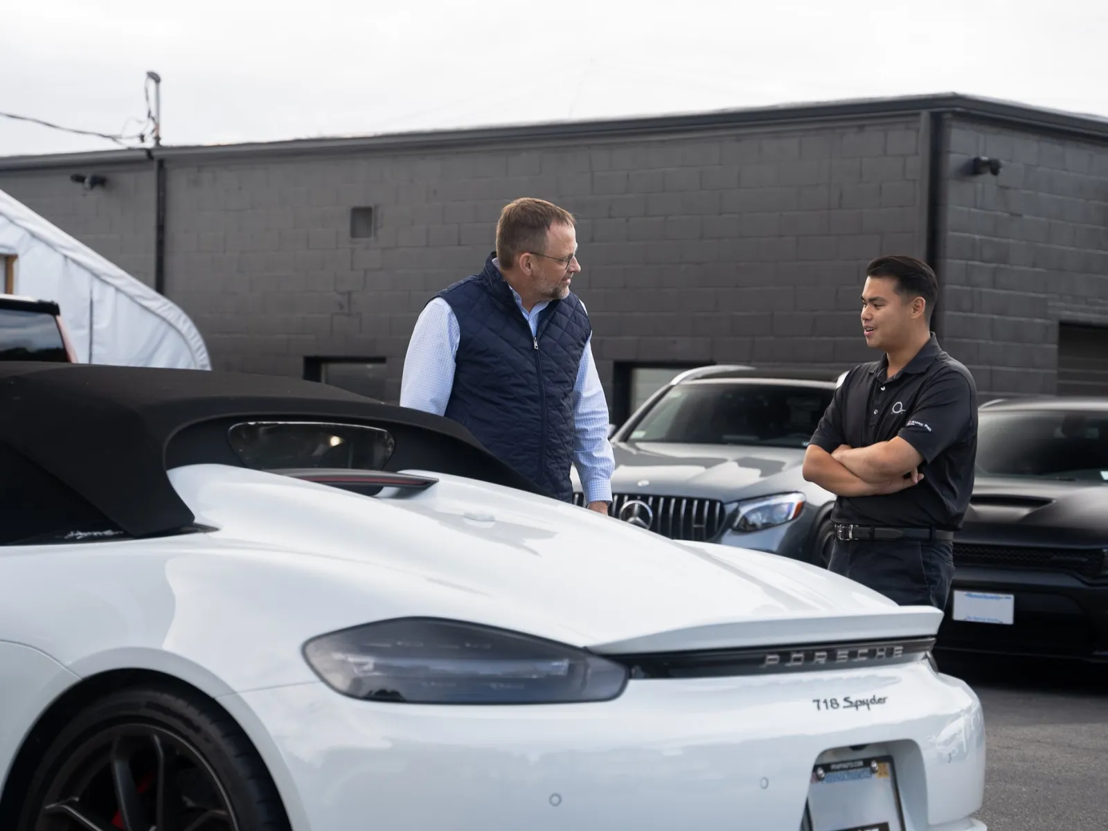 Two men talking near a white Porsche 718 Spyder convertible in a parking lot with other cars.