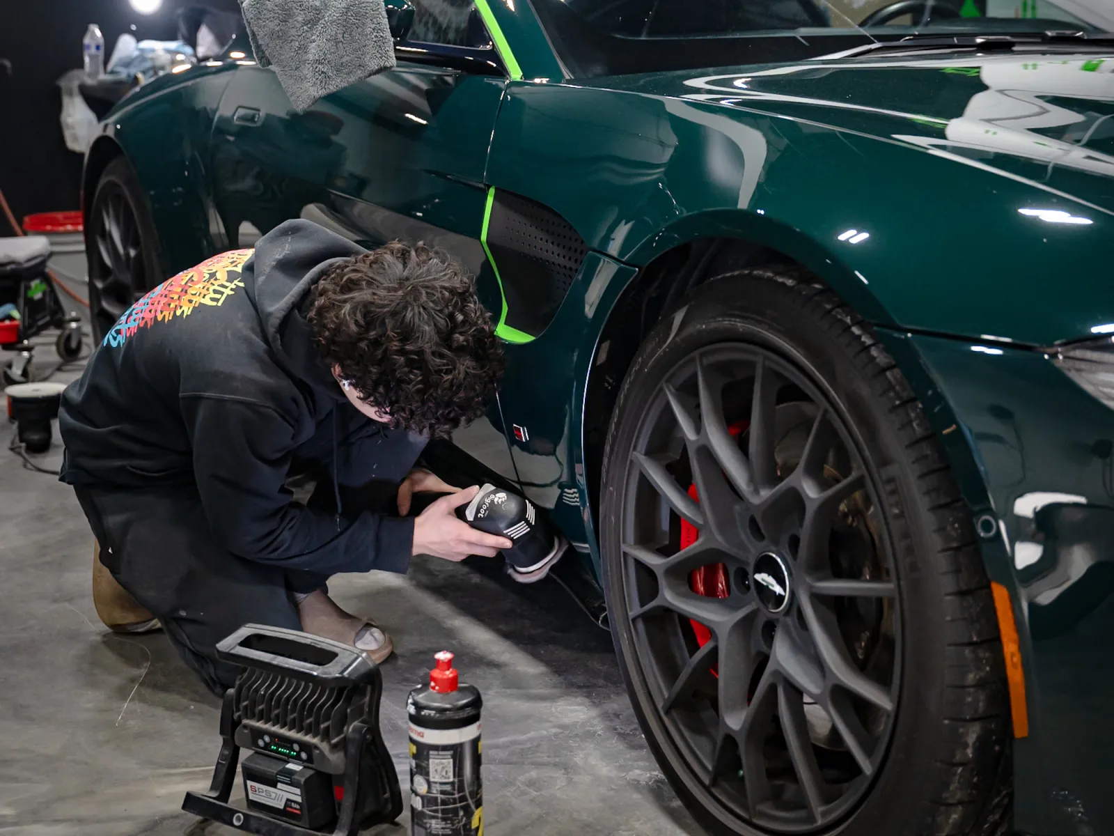 Person detailing a dark green sports car's wheel using a polishing tool in a garage with bright lights.