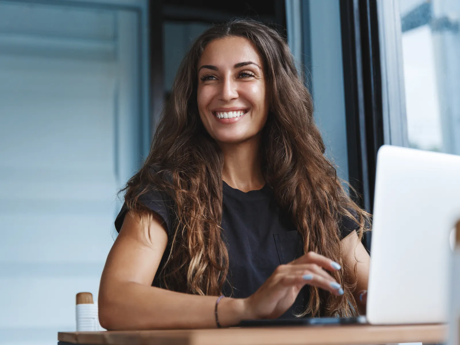 Smiling woman with long hair working on a laptop in bright modern office setting.