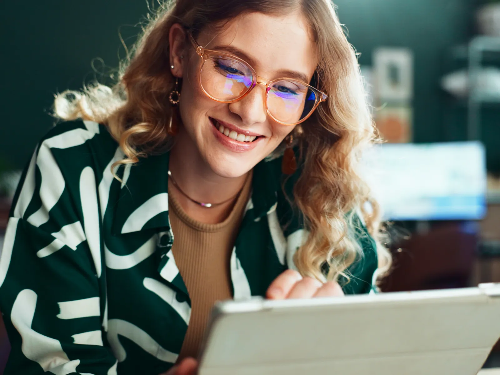 Young woman with glasses smiling while using a tablet in a cozy indoor setting with blurred background