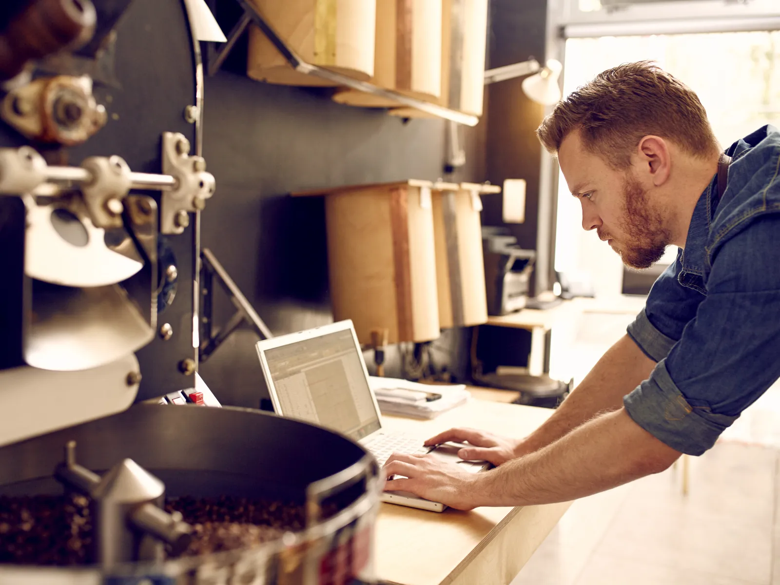 Man working on a laptop next to a coffee roasting machine with roasted coffee beans