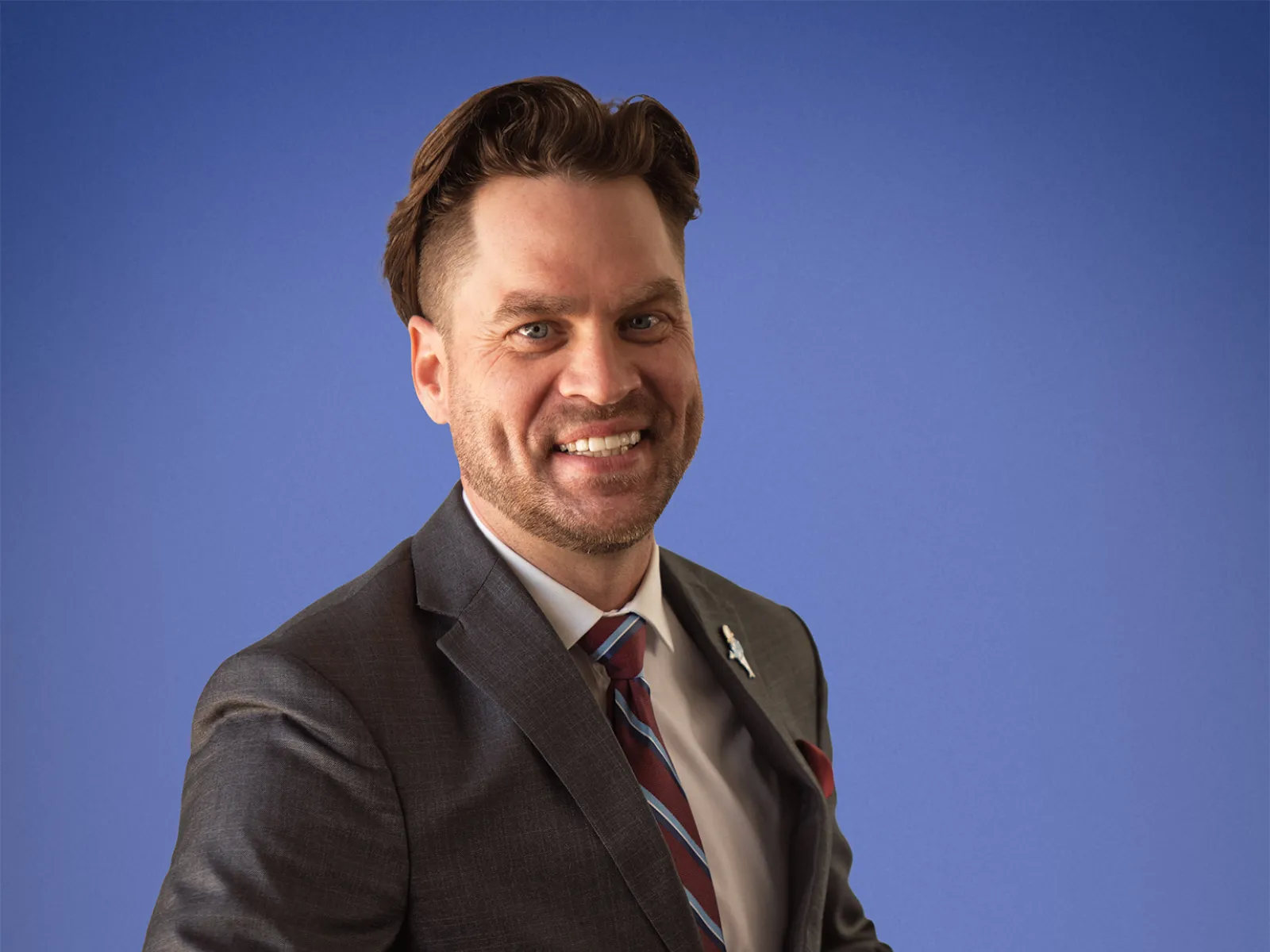 Smiling man in a dark suit adjusting jacket with a striped tie against a blue backdrop