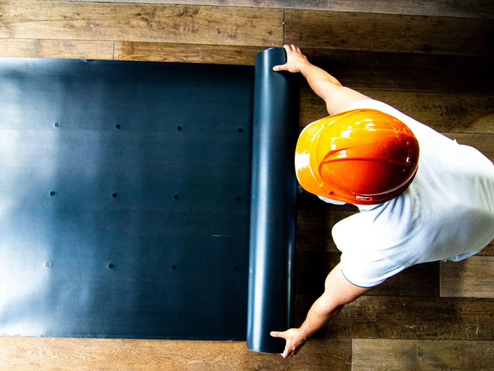 Worker wearing orange helmet unrolling black material on wooden floor for installation or construction.