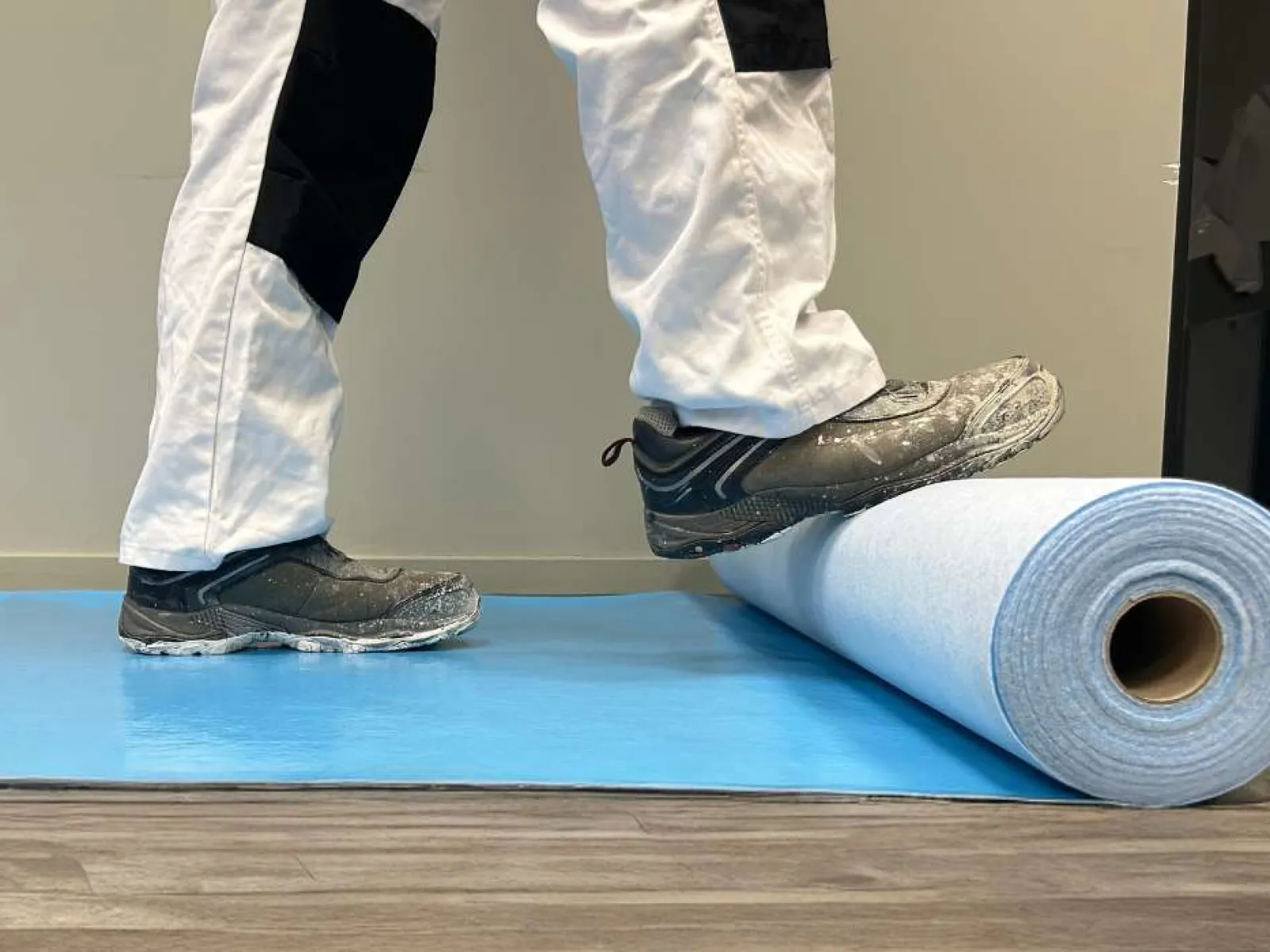 Person in work pants stepping on a rolled flooring material on a blue protective sheet over wood floor