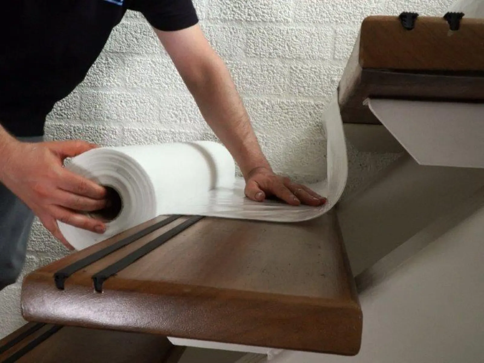 Person installing stair tread covers by unrolling anti-slip white material on wooden stairs with black safety strips.