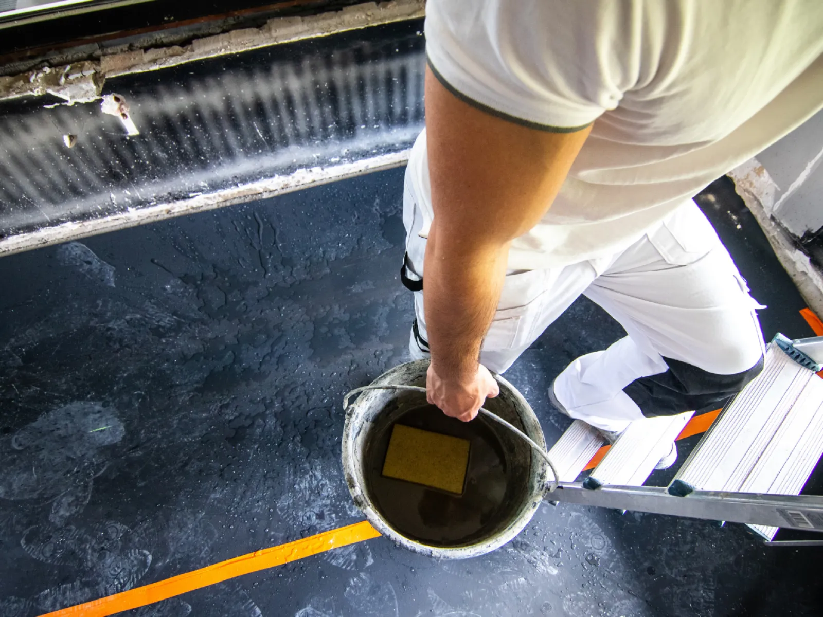 Painter in white clothes holding a bucket while standing on a ladder during interior painting work.