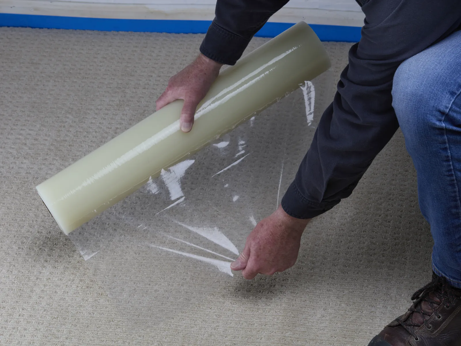 Person unrolling clear plastic protective film onto a carpeted floor to protect it from damage.